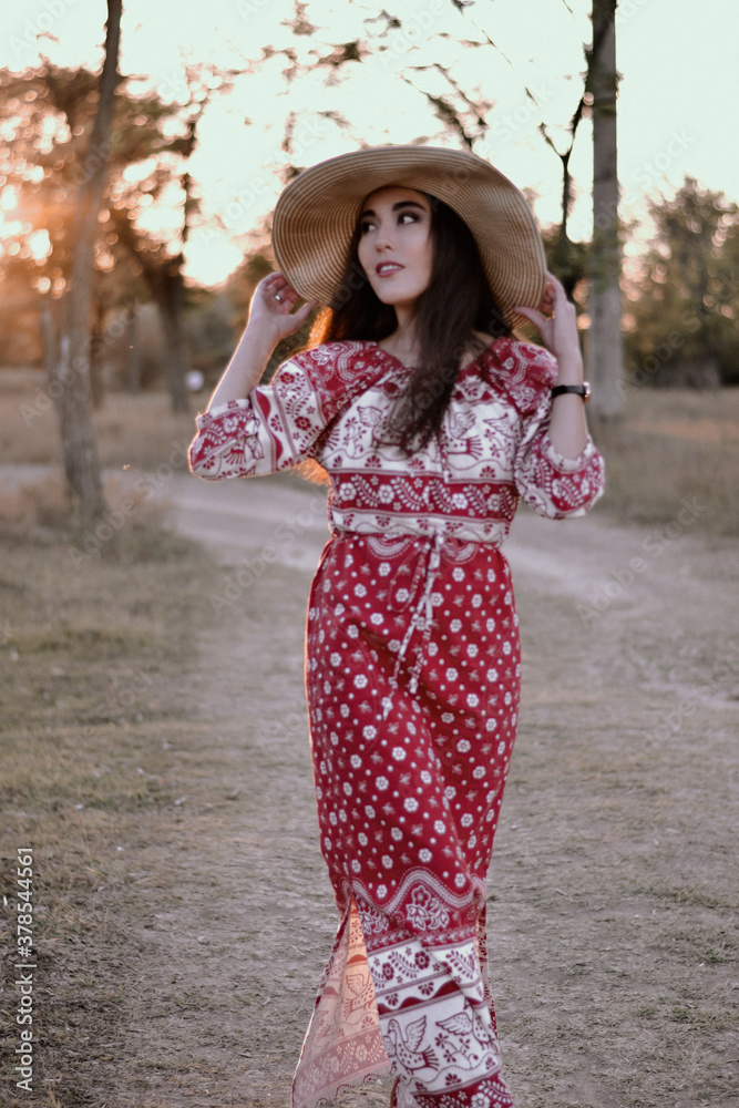 Beautiful girl with Oriental appearance in a red long sundress, hat in the Park