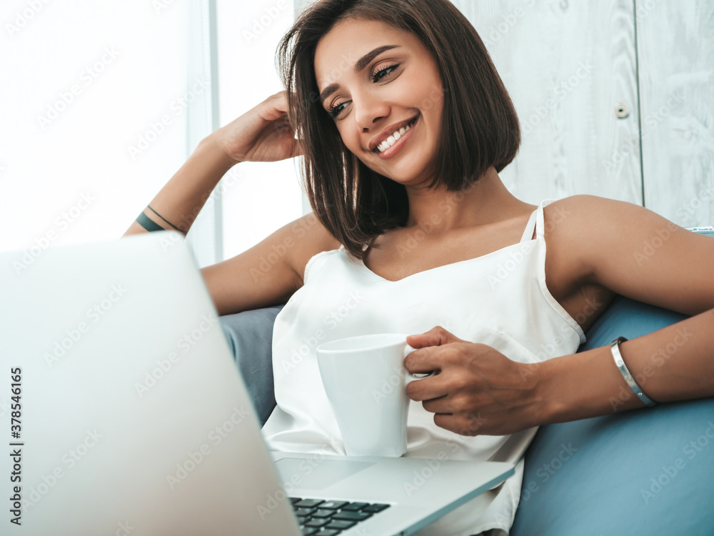 Portrait of beautiful smiling woman dressed in white pajamas. Carefree model sitting on soft bag chair and using laptop. Sexy female reading news and enjoying her morning at balcony, drinking coffee Portrait of beautiful smiling woman dressed in white pajamas. Carefree model sitting on soft bag chair and using laptop. Sexy female reading news and enjoying her morning at balcony, drinking coffee