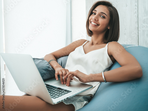 Portrait of beautiful smiling woman dressed in white pajamas. Carefree model sitting on soft bag chair and using laptop. Sexy female reading news and enjoying her morning at balcony