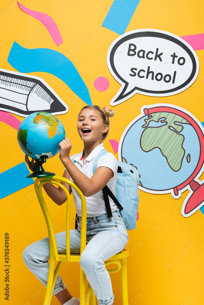 Fototapeta premium Excited schoolgirl holding globe near speech bubble with back to school lettering and paper artwork on yellow background