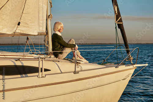 Senior relaxed woman sitting on the side of sailboat or yacht deck floating in the calm blue sea at sunset, enjoying amazing view