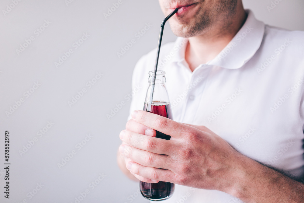 Young man in white shirt isolated over background. Cut view of guy drinking cola through plastic straw. Holding bottle in hands.