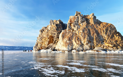 Baikal Lake in March. A group of tourists walk on the ice near the Shamanka Rock on Olkhon Island near the village of Khuzhir. Winter ice travel on the frozen lake. Beautiful landscape