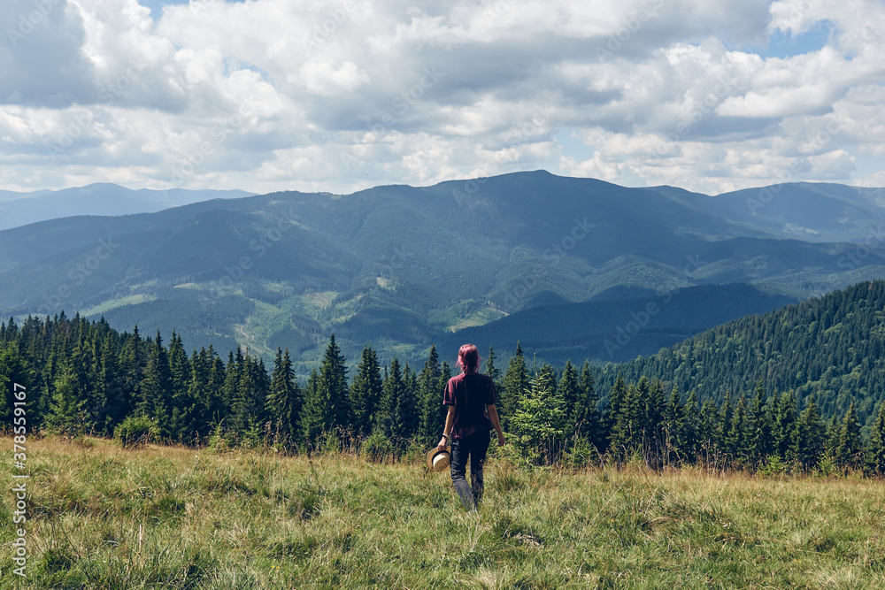 Fototapeta premium Girl with pink hair climbs uphill mountain. Hiking through forest in summer. Standing on the clearing and looks at the mountains. Dark autumn forest. Local Travel