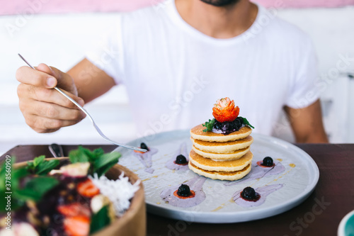 Cropped image of male holding fork sitting at dinner table eating meals for healthy diet nutrition, pancakes with sauce and strawberry fruit and bowl with ripe low calorie fruit salad for dessert