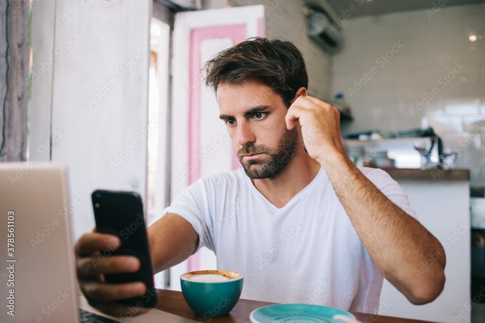 Thoughtful young bearded man using smartphone while drinking coffee in cafe Thoughtful young bearded man using smartphone while drinking coffee in cafe
