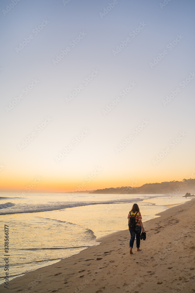 Naklejka premium Young woman walking along the golden sandy beach and fog with shoes in hand on Falesia Beach at sunset on the Atlantic Ocean. Olhos de Agua, Algarve, Portugal.