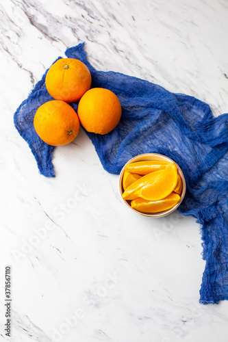 Top View of Sliced Oranges in a Blue and White Bowl on a White Countertop; Three Whole Oranges in Background with Blue Cloth photo