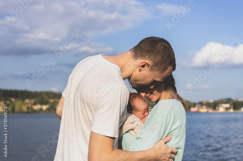 A man and a woman mom and dad carefully hug the baby between them on the background of the lake