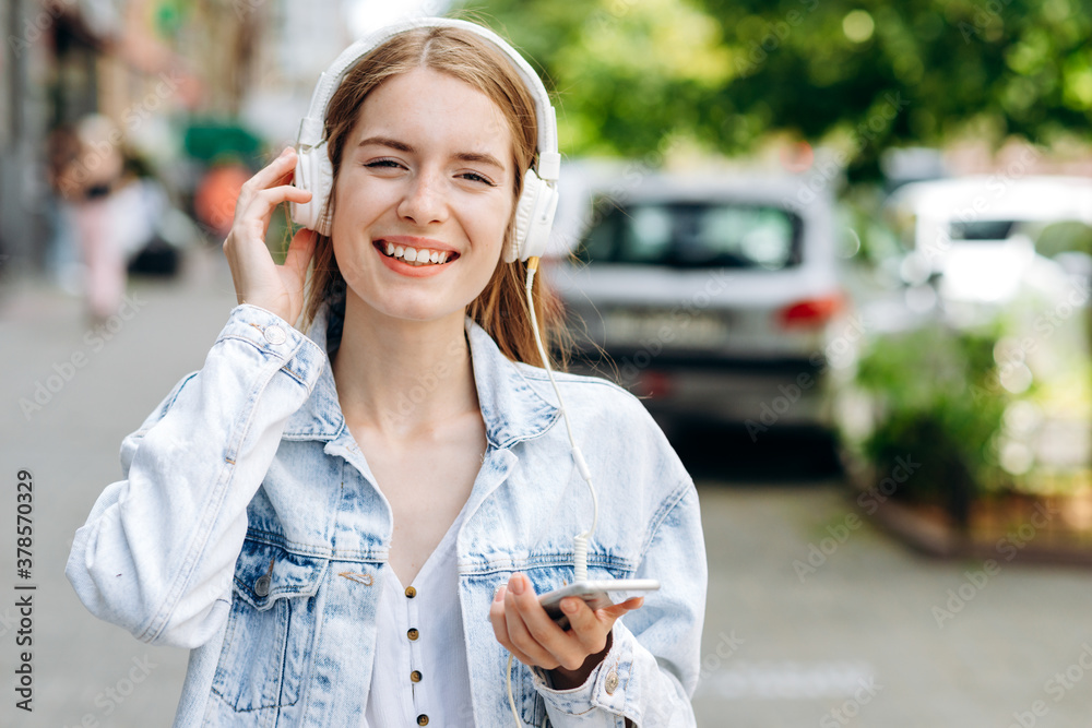 Young happy stylish trendy casual hipster woman changes songs and tracks on smartphone while listening to music on a headphone while walking around the city. Music lover enjoying music Young happy stylish trendy casual hipster woman changes songs and tracks on smartphone while listening to music on a headphone while walking around the city. Music lover enjoying music