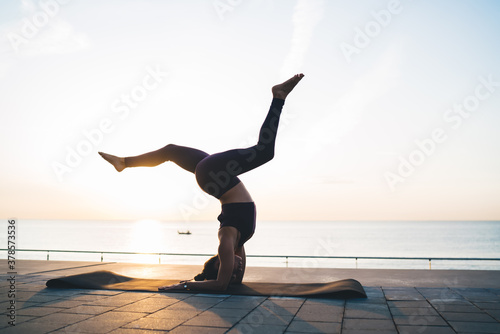 Calm woman in active wear enjoying recreation during yoga activity at embankment, strong female with athletic body standing on hands and head in asana stretching muscles reaching health and balance