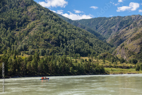 Colorful landscape of green mountains and a river with tourists floating on an inflatable boat on a summer day with blue sky and white clouds in Altai.