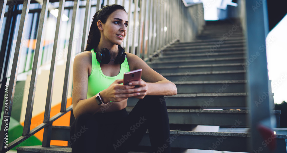 Fototapeta premium Pensive athletic woman with smartphone sitting on steps and thinking over read message
