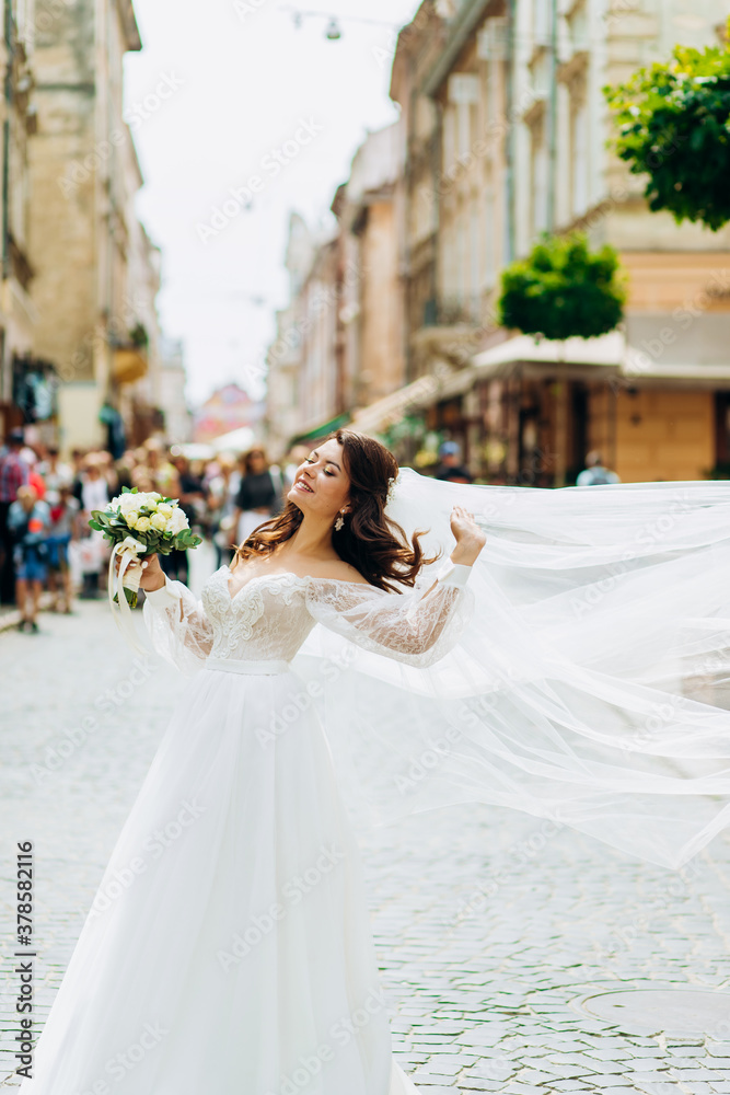 Naklejka premium Happy bride with a long veil posing on the streets of the city.