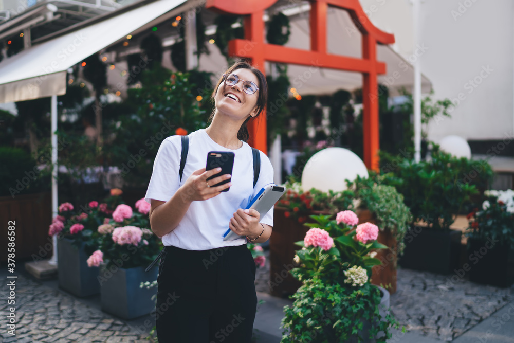 Cheerful caucasian female in casual wear holding mobile phone dialing number standing on street, smiling woman 20s millennial using mobile phone connected to 4G internet spending time in city