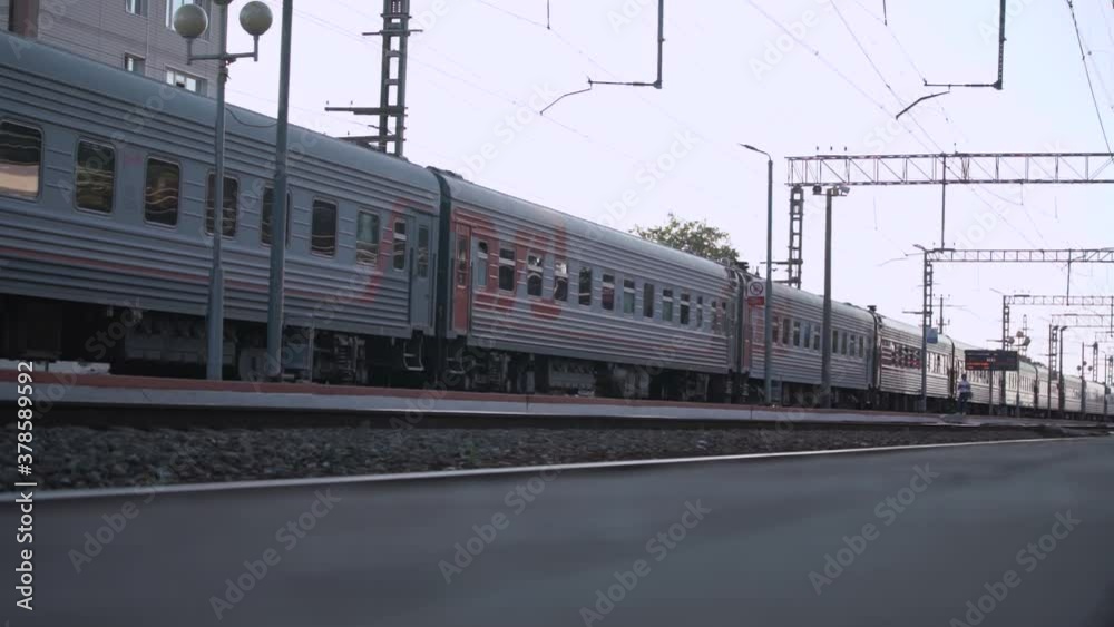 Long passenger grey train drives on rails past shady asphalt platform leaving station against city building under white sky on summer day slow motion Long passenger grey train drives on rails past shady asphalt platform leaving station against city building under white sky on summer day slow motion