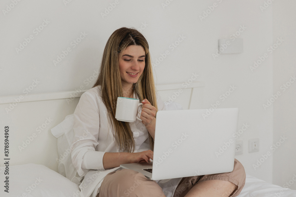 Young beautiful woman relaxing and drinking cup of hot coffee or tea using laptop computer on the bed in the bedroom. Online chat. Working from home. Woman checking social apps. Online order food Young beautiful woman relaxing and drinking cup of hot coffee or tea using laptop computer on the bed in the bedroom. Online chat. Working from home. Woman checking social apps. Online order food