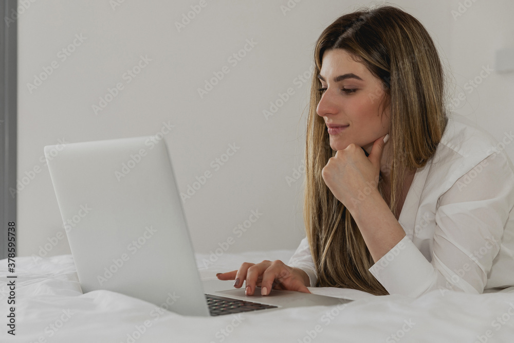 Young beautiful woman relaxing and lies on the bed using laptop computer in the bedroom. Online chat. Working from home. Woman checking social apps. Online order food. Quarantine time. Freelancer Young beautiful woman relaxing and lies on the bed using laptop computer in the bedroom. Online chat. Working from home. Woman checking social apps. Online order food. Quarantine time. Freelancer
