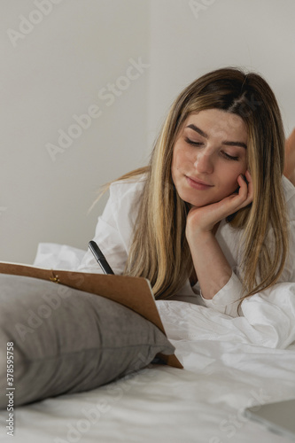 Beautiful young woman laying on bed and writing a diary in bedroom at home. Lifestyle image of girl making notes with pen and notebook and dreaming © Yelyzaveta Kras