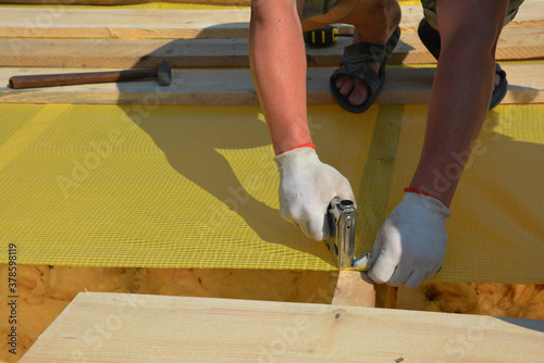 A building contractor in protective gloves is installing waterproofing vapor barrier using a staple gun while roofing construction.