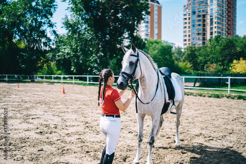 Back view of brunette professional female jockey standing near stallion horse in country club, woman rider preparing to equitation and take care for animal friend during riding vacation for resting