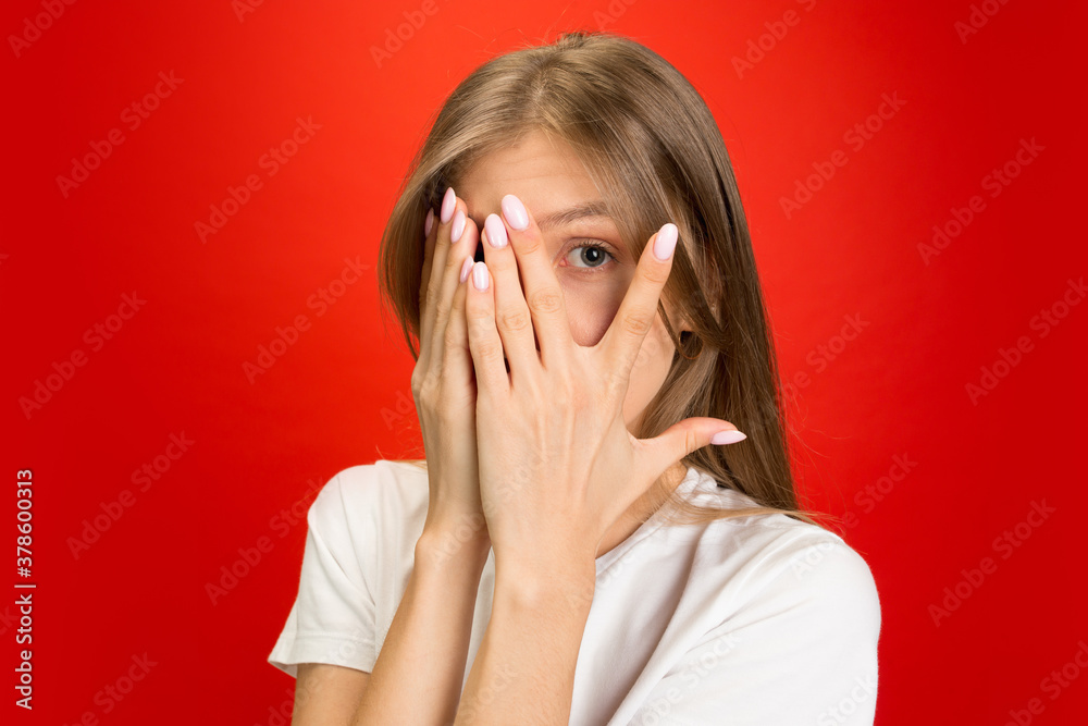 Scared hiding face. Portrait of young caucasian woman on red studio background with copyspace. Blonde female model. Concept of human emotions, facial expression, sales, advertising, youth. Scared hiding face. Portrait of young caucasian woman on red studio background with copyspace. Blonde female model. Concept of human emotions, facial expression, sales, advertising, youth.