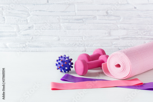 Dumbbells, fitness rubber bands, yoga mat and massage ball on white floor over white brick wall.