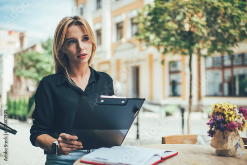 Contemplative female organisator sitting at sidewalk cafe and thinking about planning, pensive woman with paper folder pondering on information during outdoors learning and education studying photo