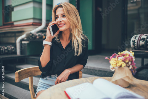 Cheerful female journalist sitting in sidewalk cafe with education textbook and using roaming internet connection on modern cellphone gadget for making international conversation with friend