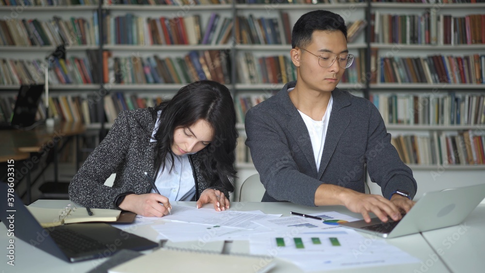 High school students working in library after classes, with paper, charts and laptops. Young man and woman sitting doing notes on background of bookshelves High school students working in library after classes, with paper, charts and laptops. Young man and woman sitting doing notes on background of bookshelves