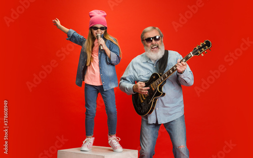 Music band. Senior man spending happy time with granddaughter on red background. Joyful elderly lifestyle, family, childhood, tech concept. Enjoying modern music, technologies. Copyspace. © master1305