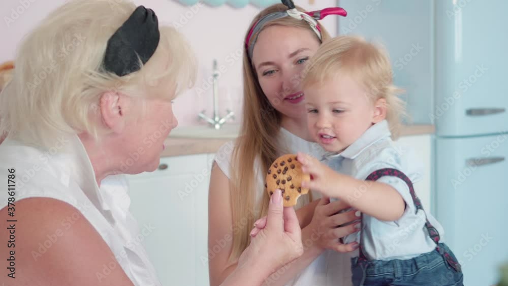 Cheerful grandma talking with active little boy and giving grandson sweet cookie. Smiling young woman admiring perfect relationship of son and mother. Portrait of happy Caucasian family indoors. Cheerful grandma talking with active little boy and giving grandson sweet cookie. Smiling young woman admiring perfect relationship of son and mother. Portrait of happy Caucasian family indoors.