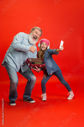 Devices of times. Senior man spending happy time with granddaughter on red background. Joyful elderly lifestyle, family, childhood, tech concept. Enjoying modern music, technologies. Copyspace. photo