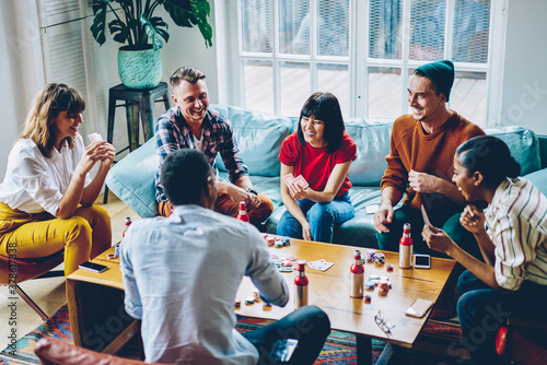 Cheerful multicultural hipster guys having fun sitting at poker table with drinks and chips, sincerely diverse people 20 years old laughing while discussing new board game in modern apartment