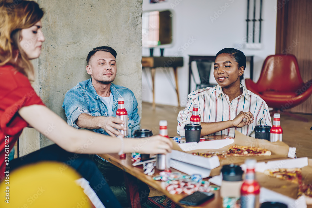 Fototapeta premium Hipster friends sitting at table with delivered pizza and alcohol drinks discussing board game with chips for playing poker during weekend, multicultural people enjoying gaming entertainment
