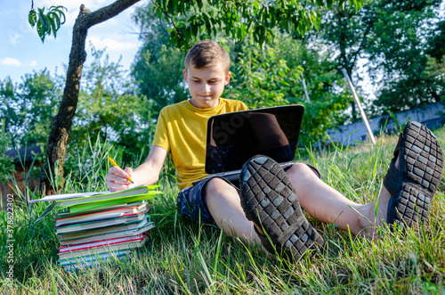 The guy is a student, a schoolboy is engaged, doing lessons online in the garden under the tree. 