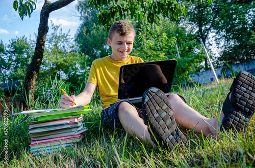 The guy is a student, a schoolboy is engaged, doing lessons online in the garden under the tree. 