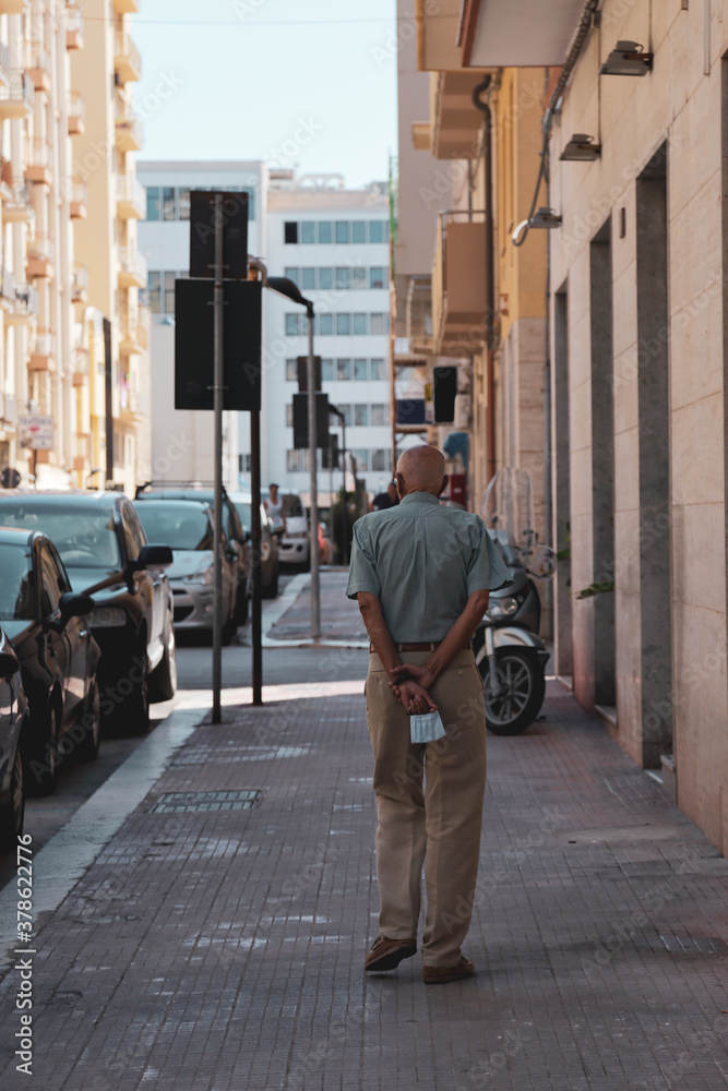Persona anziana di spalle passeggia tra le vie urbane con la mascherina in mano
