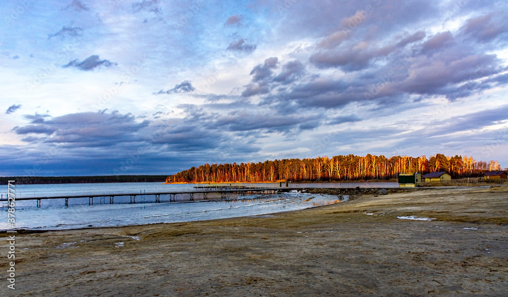 Autumn landscape on the lake. Yellow forest, cloudy sky, lake in ice and sandy beach. Natural background