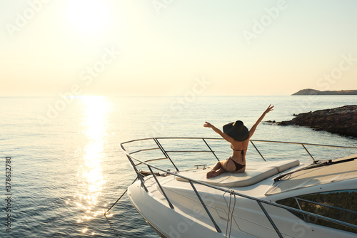 Attractive woman with big hat sitting at white luxury yacht with sea and sun track on water at background