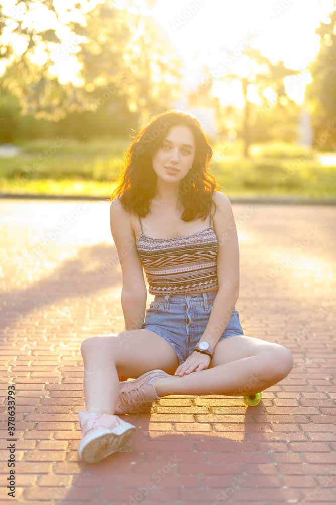 Beautiful active brunette girl with long hair wearing top, blue denim shorts and stylish pink sneakers posing on sunset background. Girl holding a orange skate with green wheels. girl sitting
