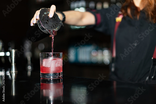 Bartender pouring cocktail from shaker into glasses at bar counter in bar, focus on coctail