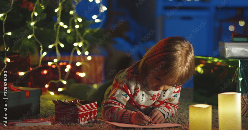 Portrait shot of the lovely small child lying on the floor and drawing a picture on Christmas Eve at the decorated x-mas tree.