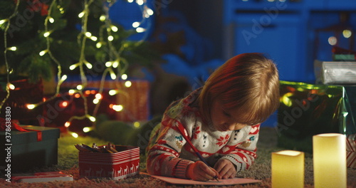 Portrait shot of the lovely small child lying on the floor and drawing a picture on Christmas Eve at the decorated x-mas tree.