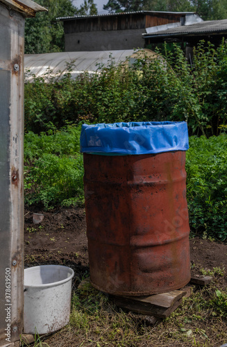 old red rusty barrel with blue bag inside stands in a green garden among greenhouses