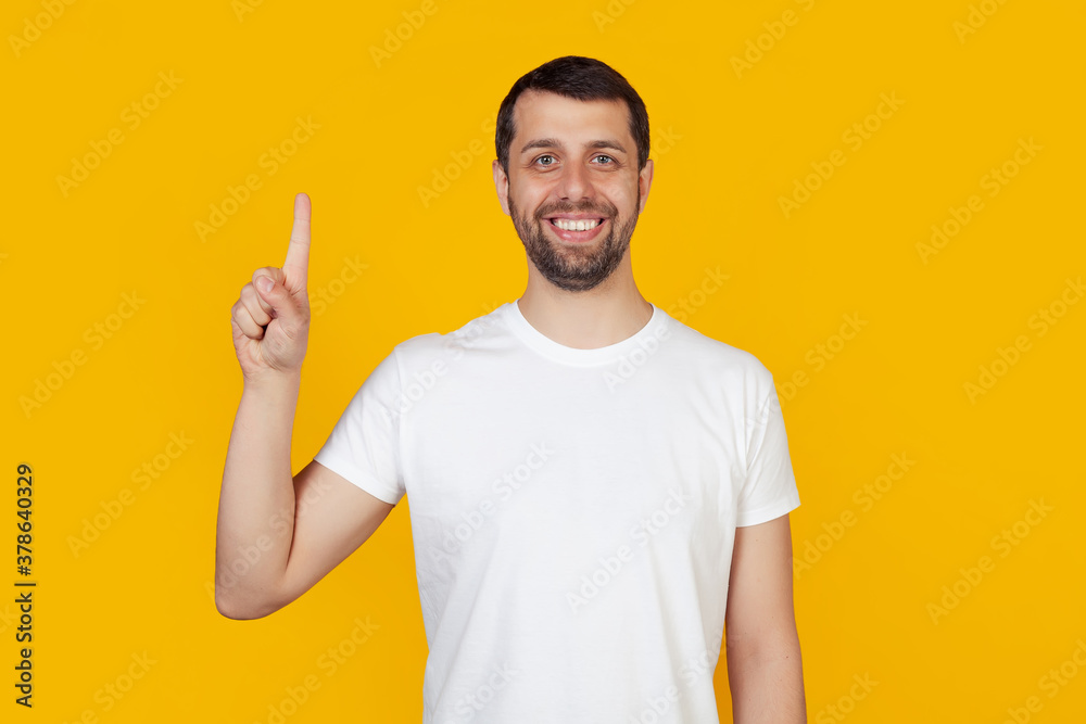 Modern young man with a beard in a white tank top shows number one with fingers on hand, smiling confidently and happily, looking at the camera. The man shows one finger. Number 1, yellow background. Modern young man with a beard in a white tank top shows number one with fingers on hand, smiling confidently and happily, looking at the camera. The man shows one finger. Number 1, yellow background.