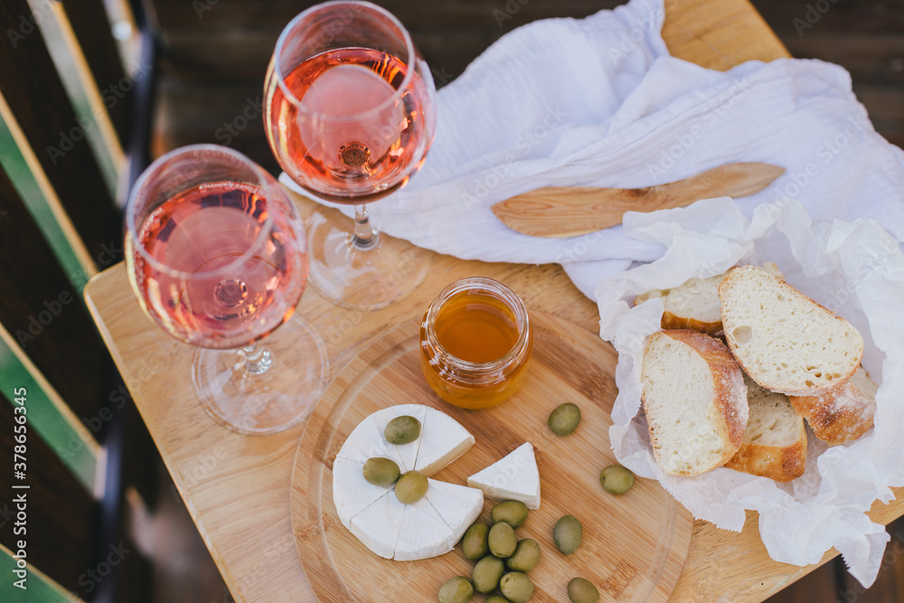 Fototapeta premium Two glasses of rose wine, cheese, olives, honey and fresh bread on balcony of a cozy wooden house with beautiful view.