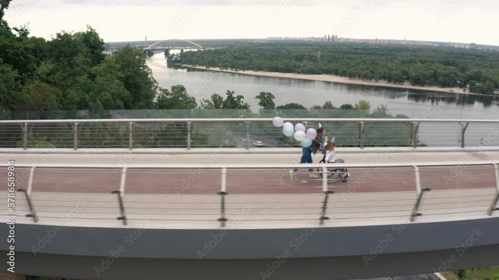 Aerial shot of excited teen daughter with paralyzed mother in cone hats having fun on birthday celebration while riding wheelchair with balloons across footbridge, joy, happiness, festive mood Aerial shot of excited teen daughter with paralyzed mother in cone hats having fun on birthday celebration while riding wheelchair with balloons across footbridge, joy, happiness, festive mood