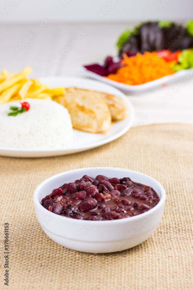 Black beans on white plate on raw cotton dish towel with a full plate with rice chicken and fries on background. feijoada Black beans on white plate on raw cotton dish towel with a full plate with rice chicken and fries on background. feijoada