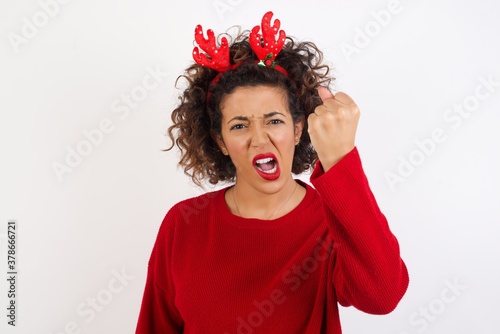 Young arab woman with curly hair wearing christmas headband standing on white background being angry and mad raising fist frustrated and furious while shouting with anger. Rage and aggressive concept.